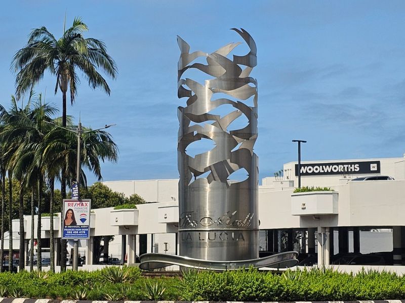 A view of a landscaped roundabout featuring a prominent metal sculpture, situated in front of a commercial shopping center.