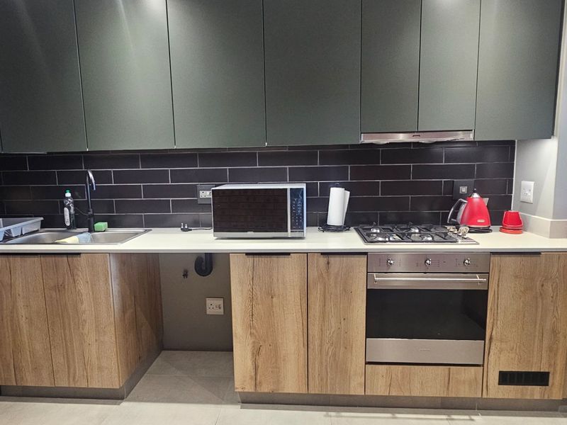 Modern kitchen featuring dark matte cabinetry, wood-grain lower cabinets, subway tile backsplash, gas hob, built-in oven, and stone countertops.