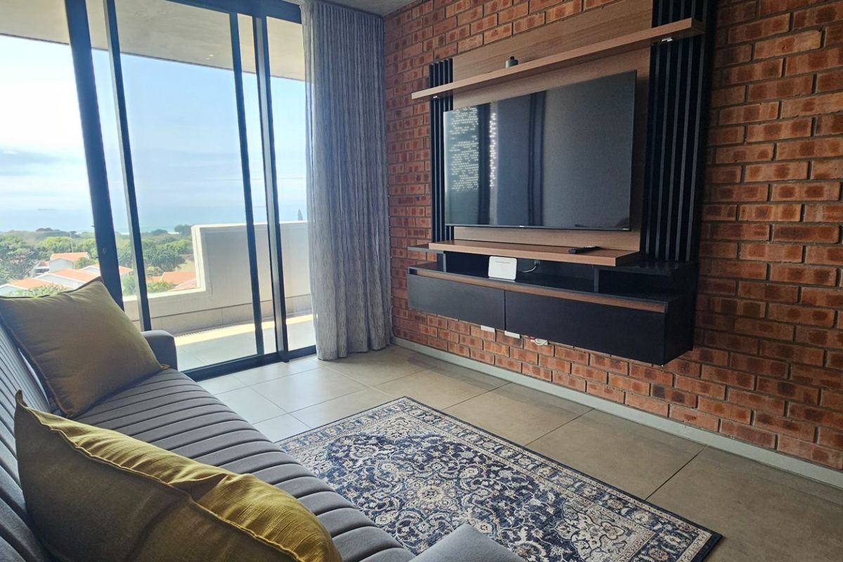 A modern living room featuring an exposed brick wall, built-in media console, and large sliding glass doors leading to a balcony with coastal views.