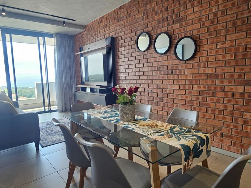 Modern open-plan living and dining area featuring an exposed brick wall, tiled floors, and large sliding doors leading to a balcony with scenic views.