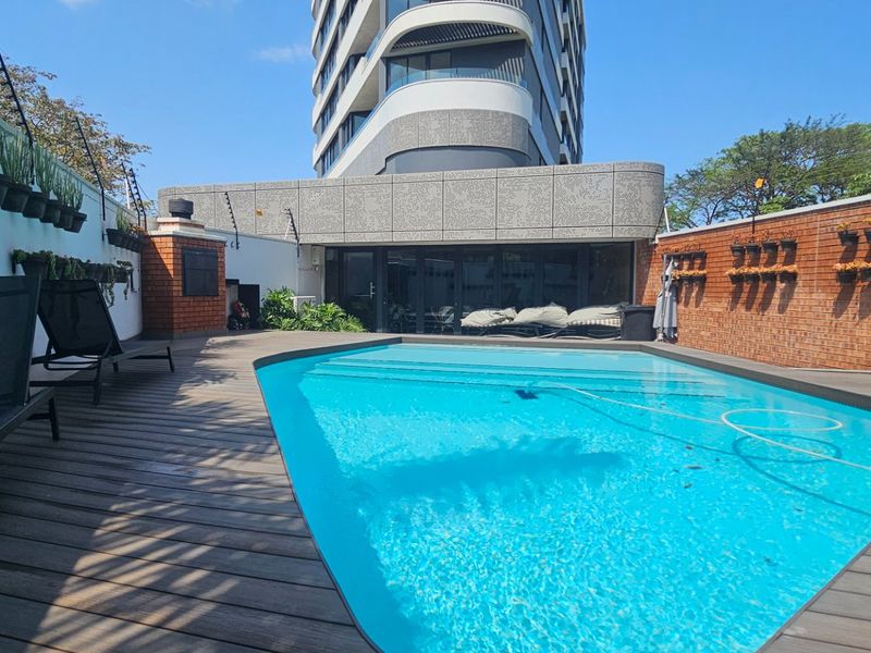 A well-maintained outdoor pool area featuring timber decking, a brick boundary wall with electric fencing, and a built-in braai facility.