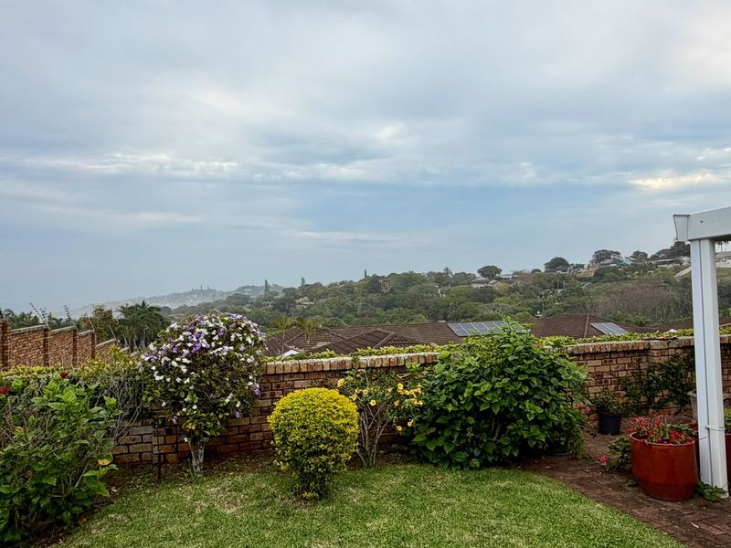 Elevated outdoor view overlooking the surrounding neighborhood and landscape, featuring a brick boundary wall, paved patio area, and established garden.