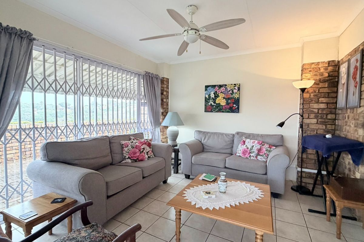 A comfortable living room featuring tiled flooring, an exposed brick accent wall, and a ceiling fan, with access to an outdoor area via security gates.