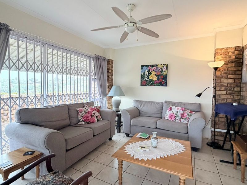 A comfortable living room featuring tiled flooring, an exposed brick accent wall, and a ceiling fan, with access to an outdoor area via security gates.