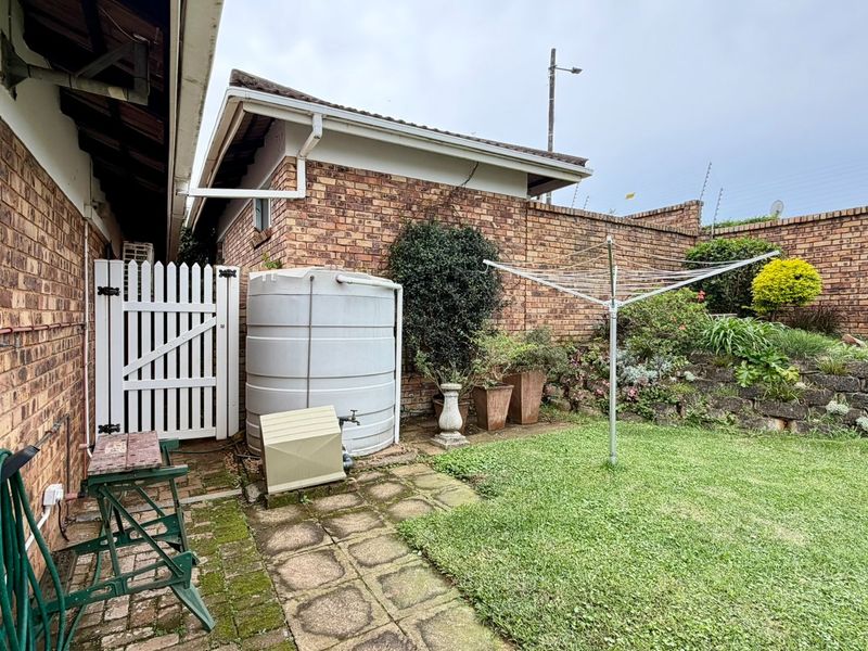 A rear exterior view of the townhouse featuring a small lawn, paved walkway, water tank, and brick boundary walls with electric fencing.