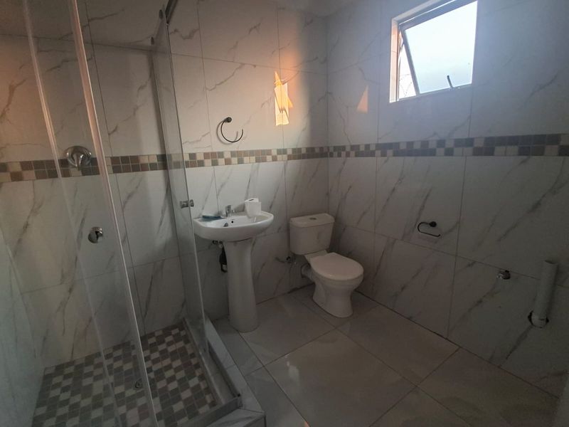 A clean, modern bathroom featuring a glass-enclosed shower, pedestal basin, and toilet with neutral marble-look wall tiles and matching floor tiles.