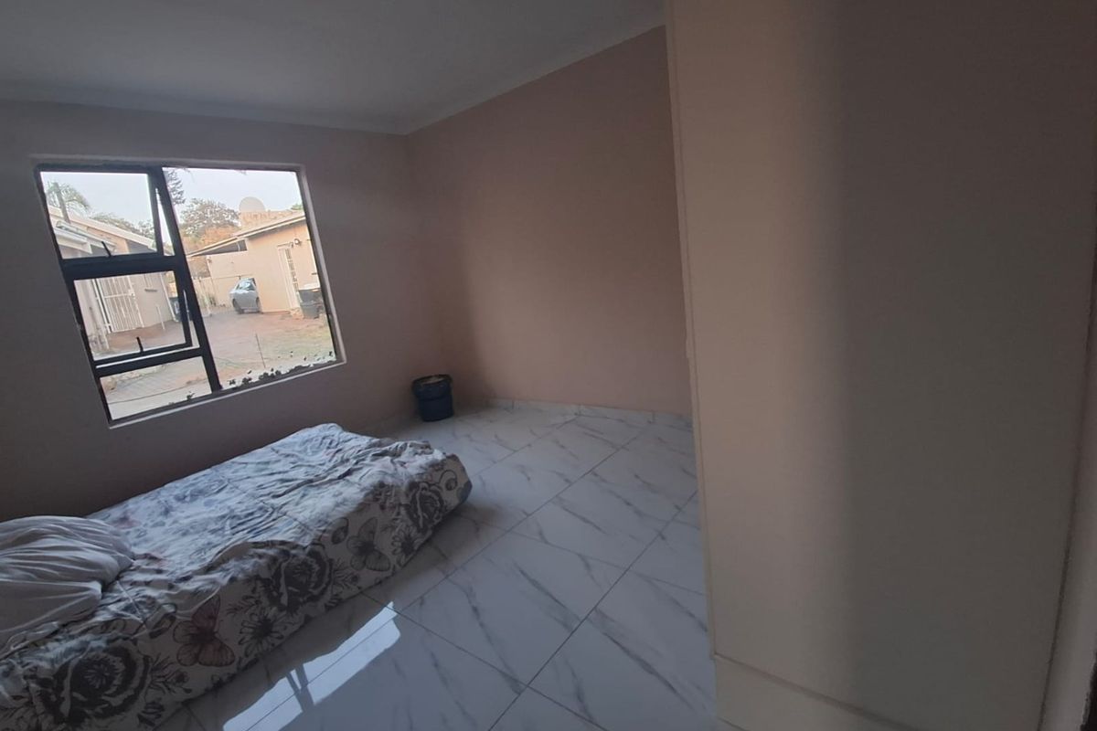 A sparsely furnished bedroom featuring white marble-look floor tiles and an aluminium window frame with a broken pane.