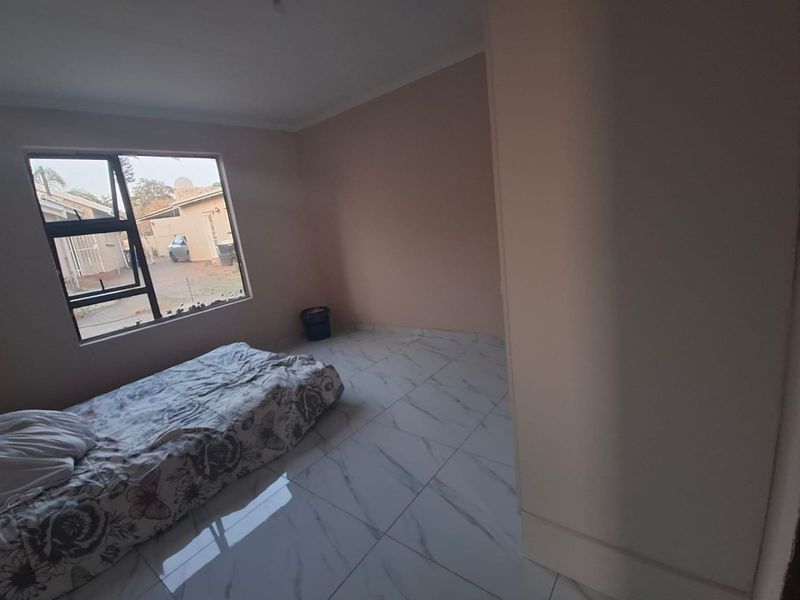 A sparsely furnished bedroom featuring white marble-look floor tiles and an aluminium window frame with a broken pane.
