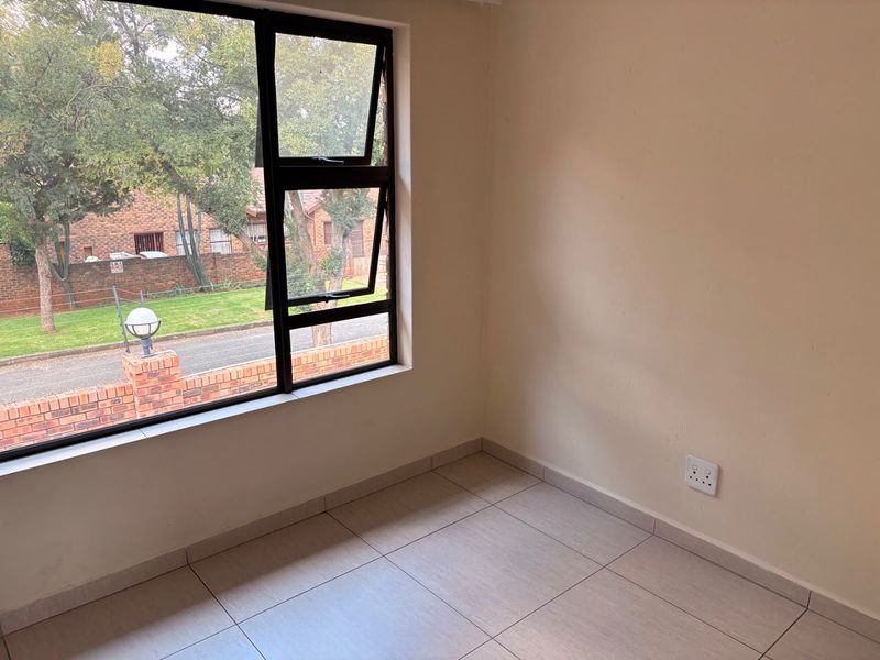 A bright, empty bedroom featuring neutral tiled flooring and a large aluminium-framed window with a view of the complex grounds.
