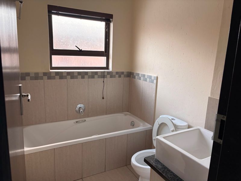 A functional bathroom featuring a bathtub, toilet, and vanity basin with neutral wall tiles and a window for natural light.