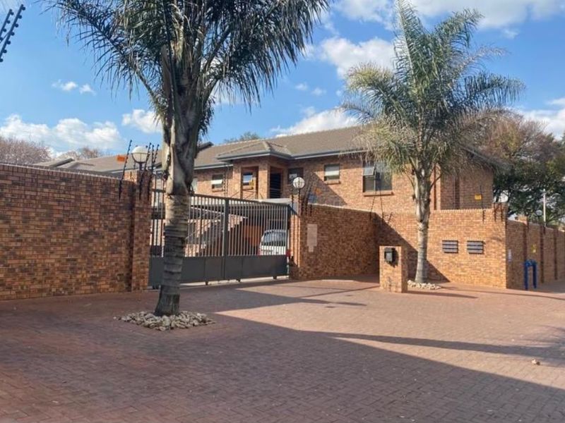 Front exterior view of a face brick apartment complex featuring a paved driveway, secure metal gate, and perimeter electric fencing.