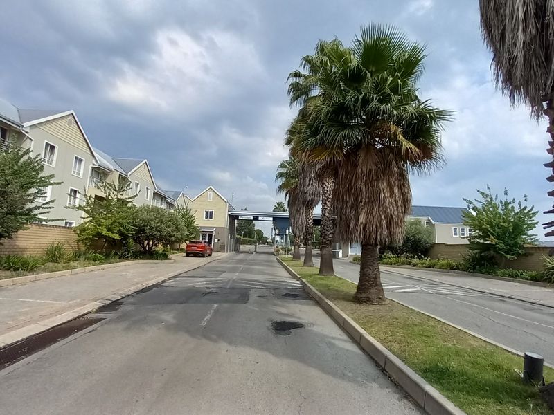 A view of the residential complex entrance road featuring mature palm trees, paved curbs, and a security gatehouse.