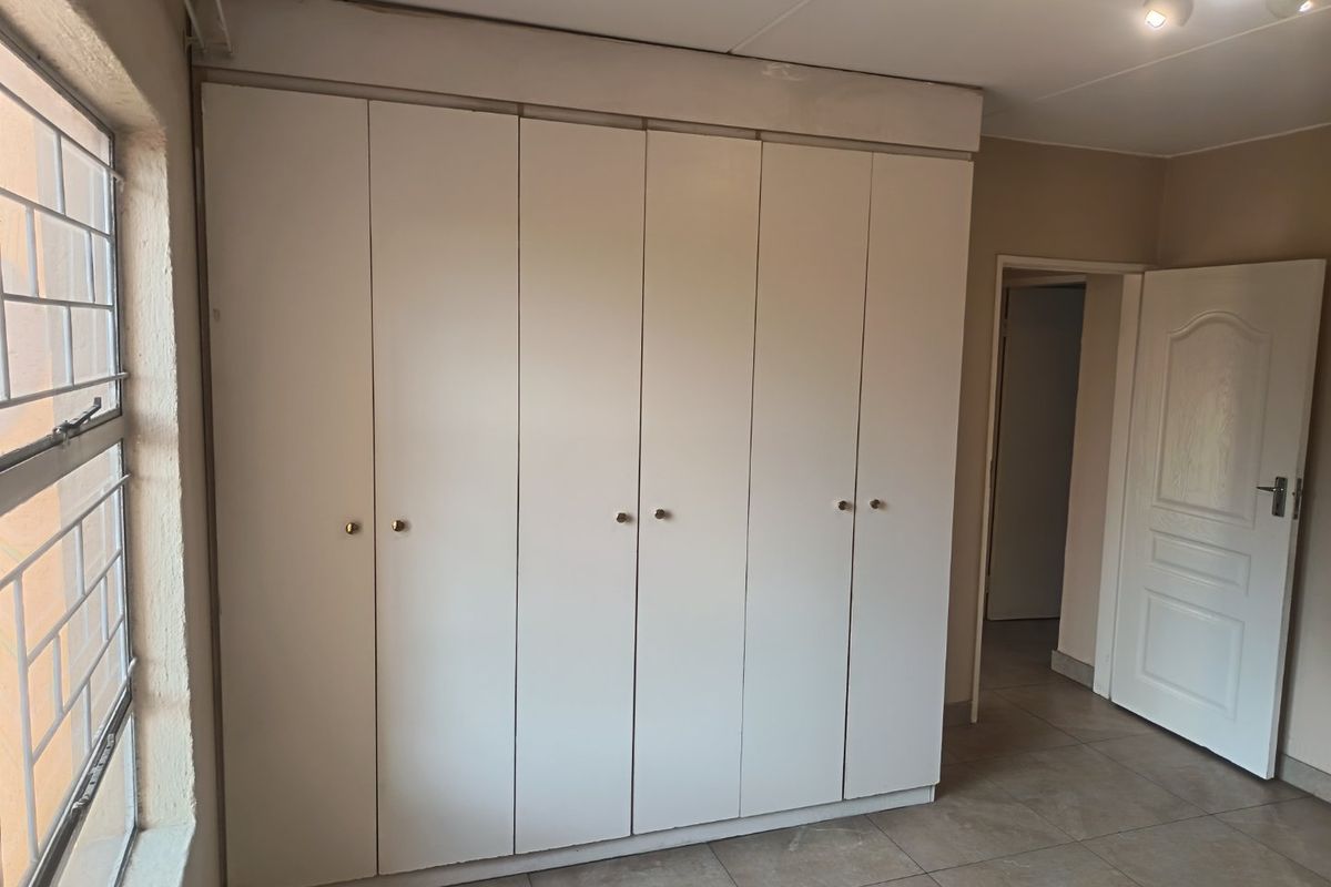 A vacant bedroom featuring neutral tiled flooring, large built-in cupboards, and a window fitted with security bars.