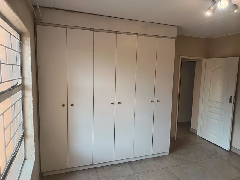 A vacant bedroom featuring neutral tiled flooring, large built-in cupboards, and a window fitted with security bars.