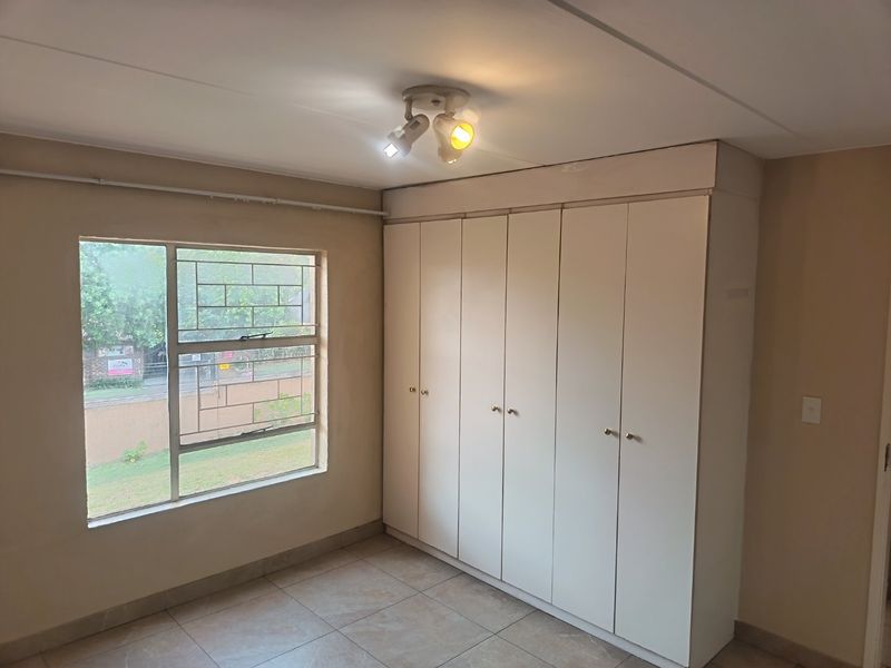 A bright, empty bedroom featuring neutral tiled flooring, built-in white cupboards, and a window with security bars.