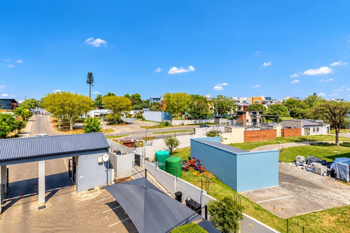 An elevated aerial view of the apartment complex entrance, featuring a security gatehouse, paved parking areas with shade cloth carports, and perimeter boundary walls.