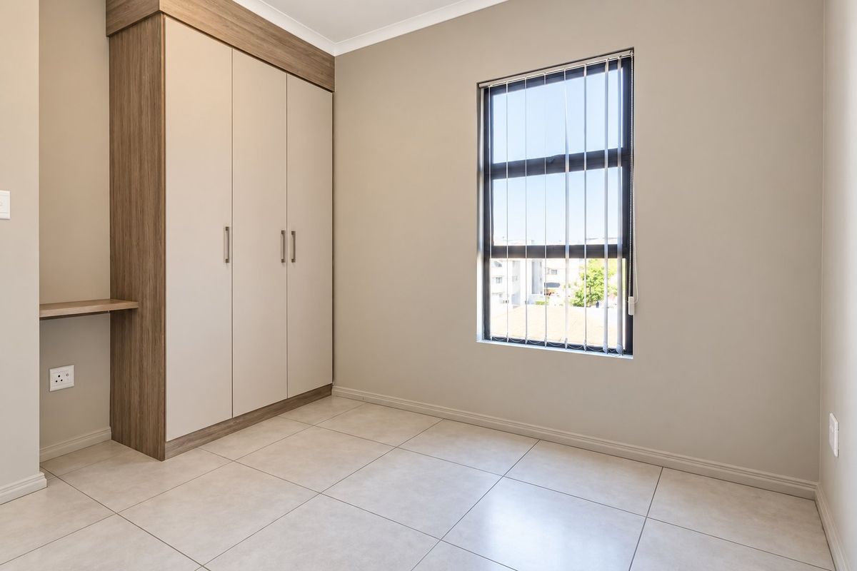A clean, modern bedroom featuring neutral tiled flooring, built-in cupboards with an integrated desk, and a window with vertical blinds.