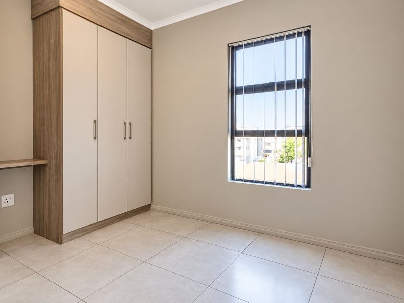 A clean, modern bedroom featuring neutral tiled flooring, built-in cupboards with an integrated desk, and a window with vertical blinds.