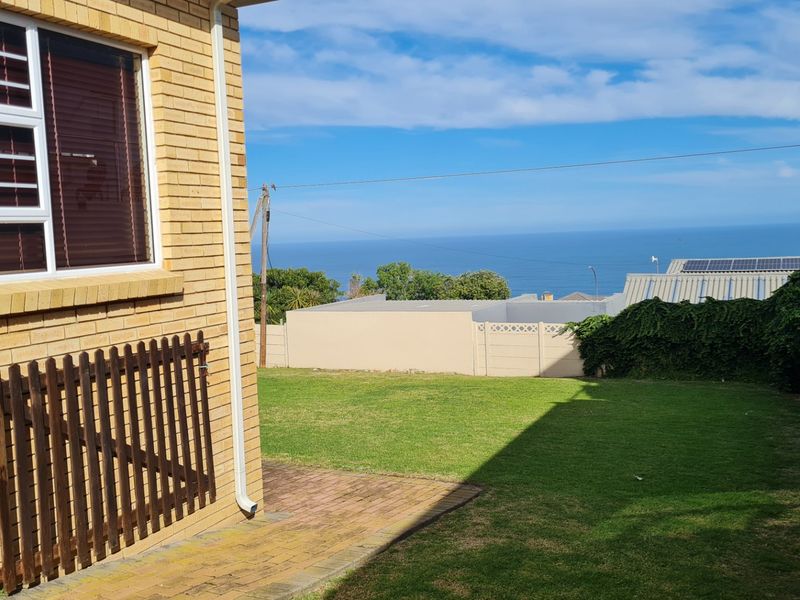 A rear exterior view featuring a brick facade, a paved patio area, a well-maintained lawn, and a boundary wall with a scenic ocean view.