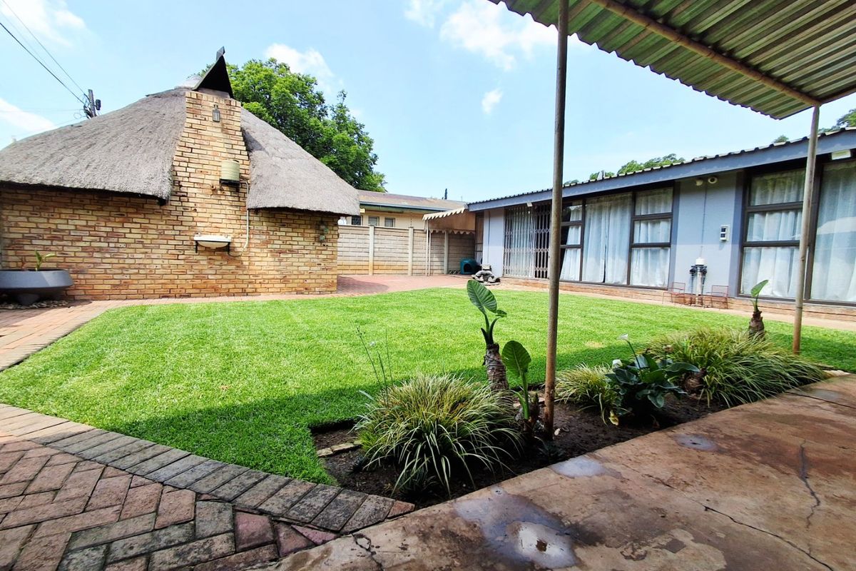 Spacious rear garden featuring a lawn, paved patio area, covered veranda, and a detached structure with a traditional thatched roof and brick chimney.