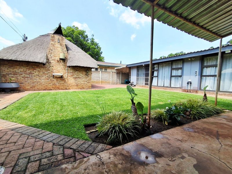 Spacious rear garden featuring a lawn, paved patio area, covered veranda, and a detached structure with a traditional thatched roof and brick chimney.