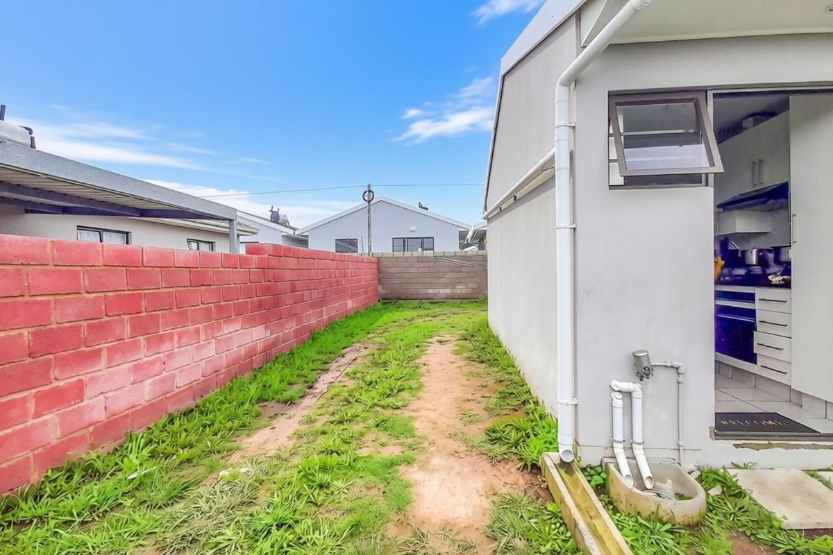 Rear exterior view showing a side pathway, brick boundary wall, and external plumbing fixtures leading to a kitchen entrance.