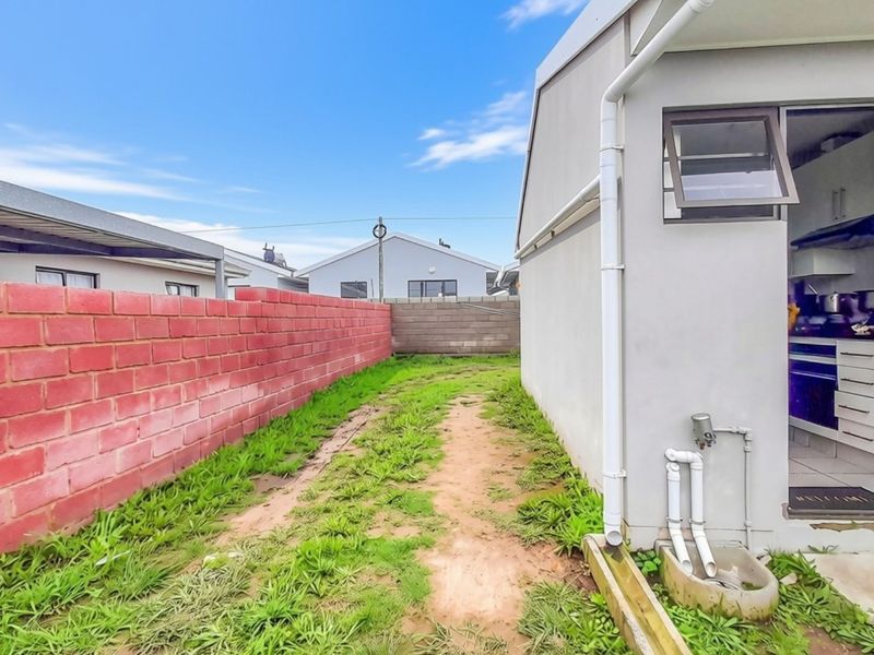 Rear exterior view showing a side pathway, brick boundary wall, and external plumbing fixtures leading to a kitchen entrance.