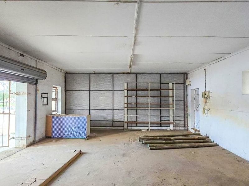 A spacious, utilitarian commercial area featuring a roller shutter door, security mesh partitioning, and concrete flooring, suitable for storage or workshop use.