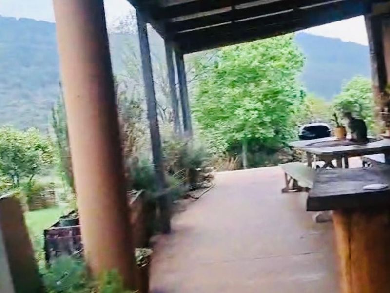 A covered outdoor patio area featuring structural columns and paved flooring, overlooking a lush garden and mountain landscape.