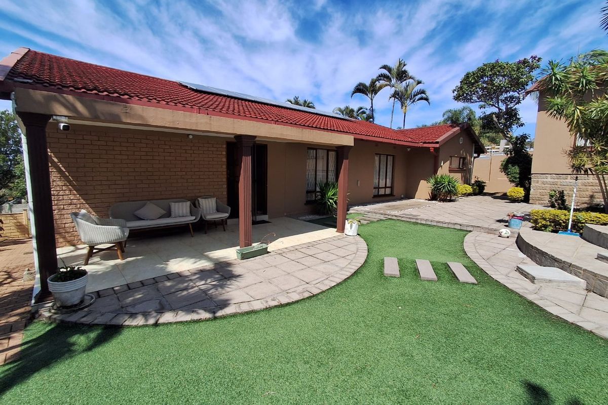 Rear view of the property featuring a covered patio, paved courtyard area, and well-maintained artificial lawn.