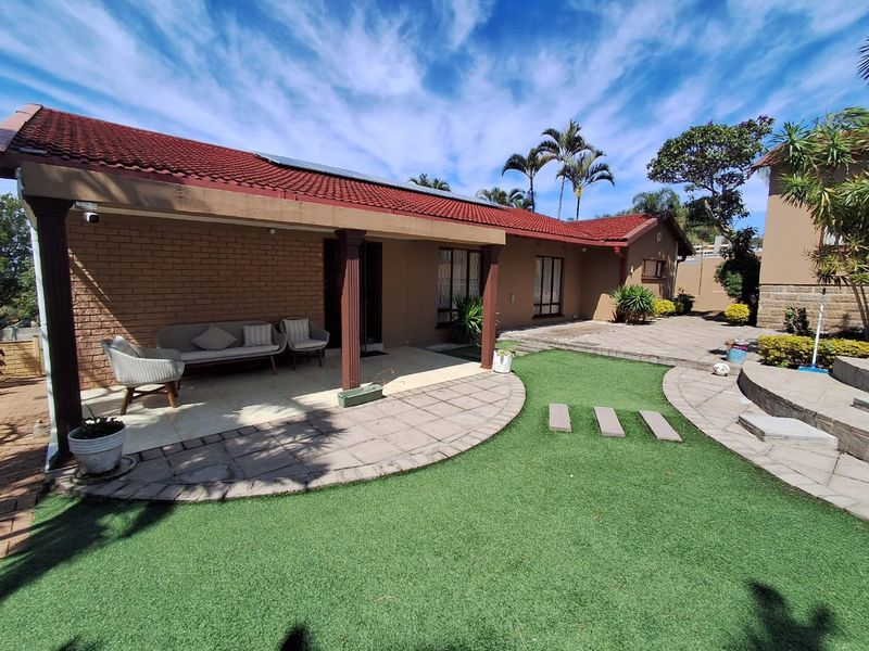 Rear view of the property featuring a covered patio, paved courtyard area, and well-maintained artificial lawn.