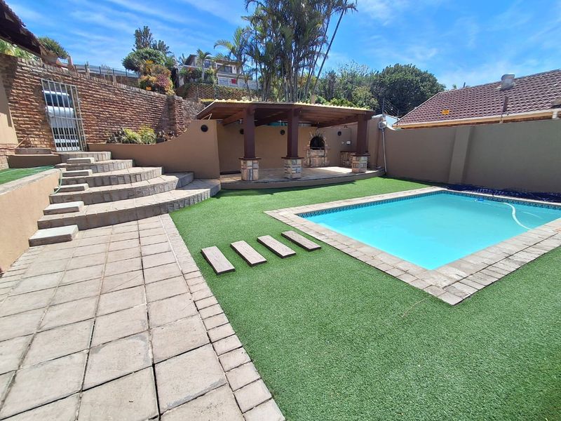 Outdoor entertainment area featuring a swimming pool, artificial lawn, paved stairs, and a covered patio with a built-in braai.