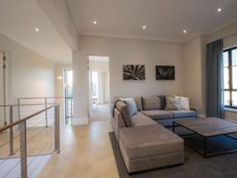 A well-lit living room featuring timber flooring, recessed downlights, and a modern metal balustrade overlooking the staircase.