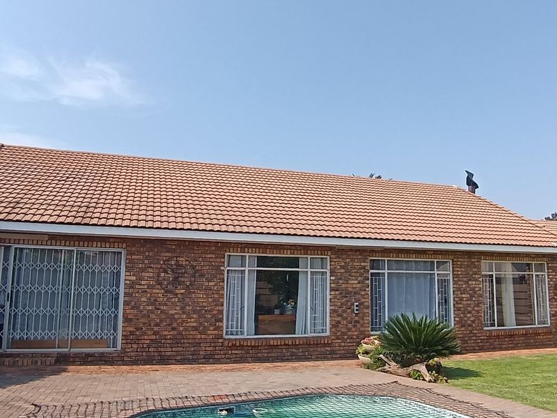 Rear view of a brick house featuring a tiled roof, large windows, security gates, a paved patio area, and a swimming pool.