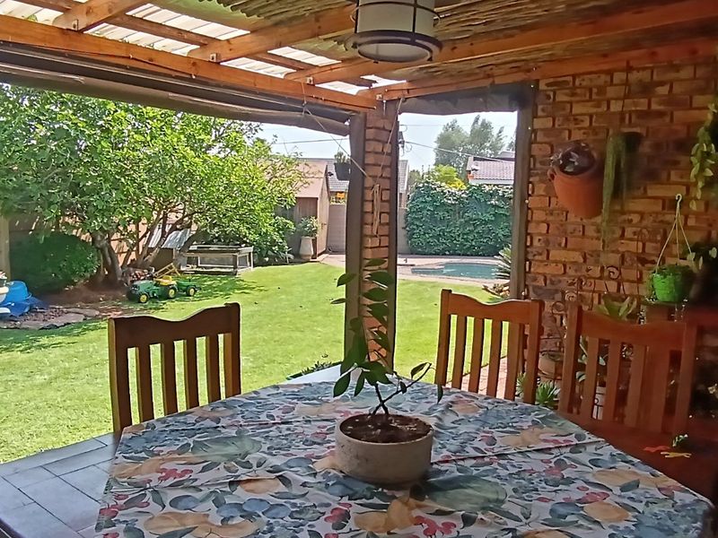 A covered outdoor patio area featuring exposed brick walls and a timber pergola roof, overlooking a private garden and swimming pool.