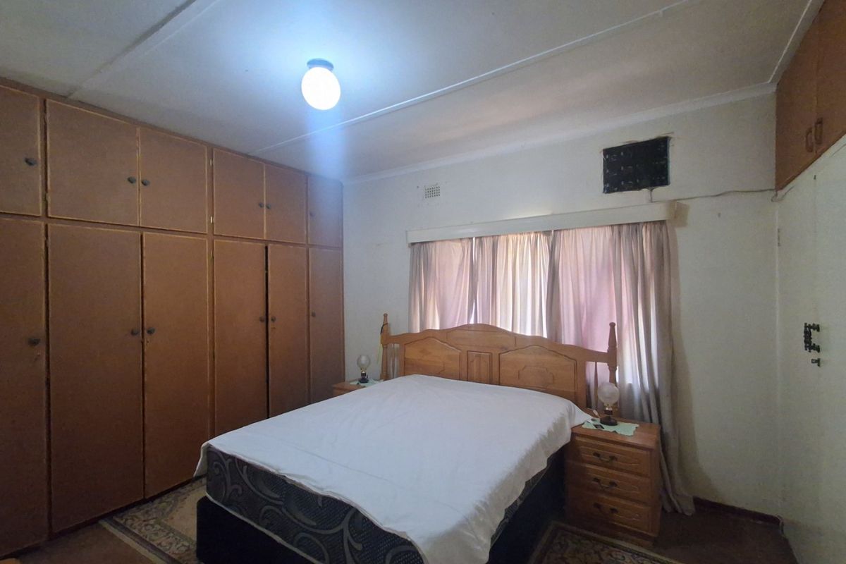 A bedroom featuring extensive built-in wooden cupboards, a central light fixture, and a window with curtains, offering a functional space with dated finishes.
