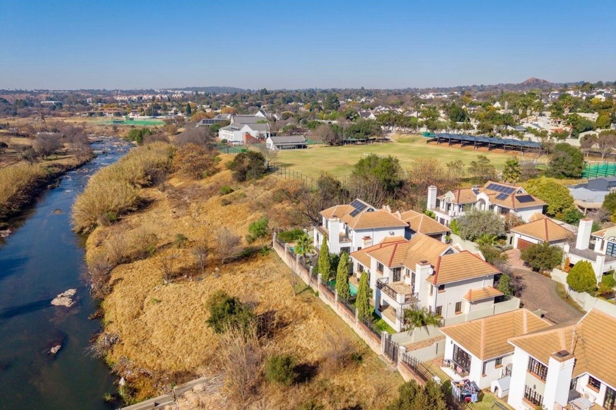 Aerial view of a townhouse complex situated along a riverbank, featuring tiled roofs, solar installations, and proximity to local sports fields.