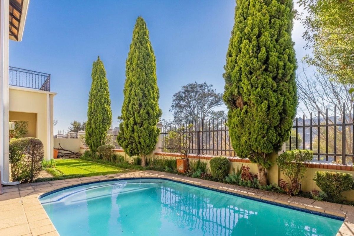 A well-maintained outdoor pool area featuring a paved surround, established cypress trees, and secure perimeter fencing with a view of the upper balcony.
