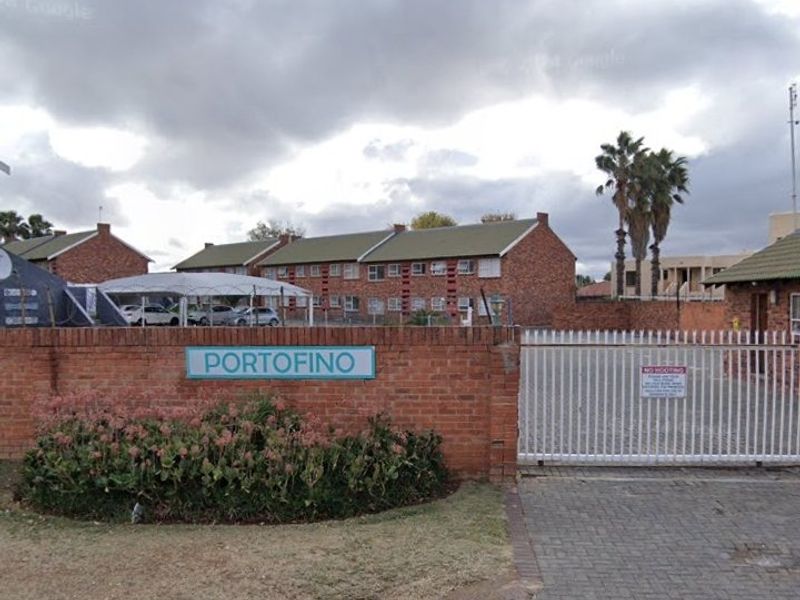 Front entrance view of the Portofino townhouse complex, featuring a brick boundary wall, secure metal gate, and paved driveway access.