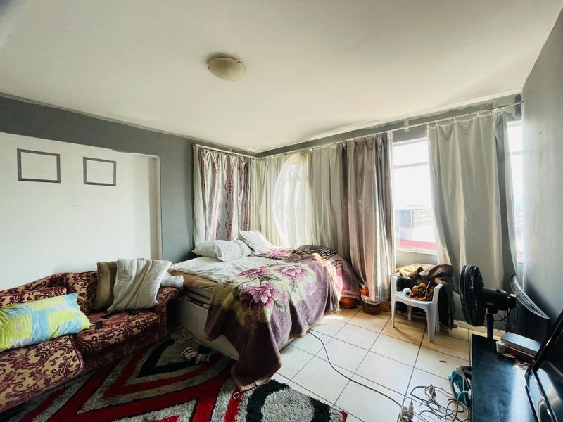 A bedroom featuring tiled flooring and large windows with curtains, currently used as a multi-functional living and sleeping space.