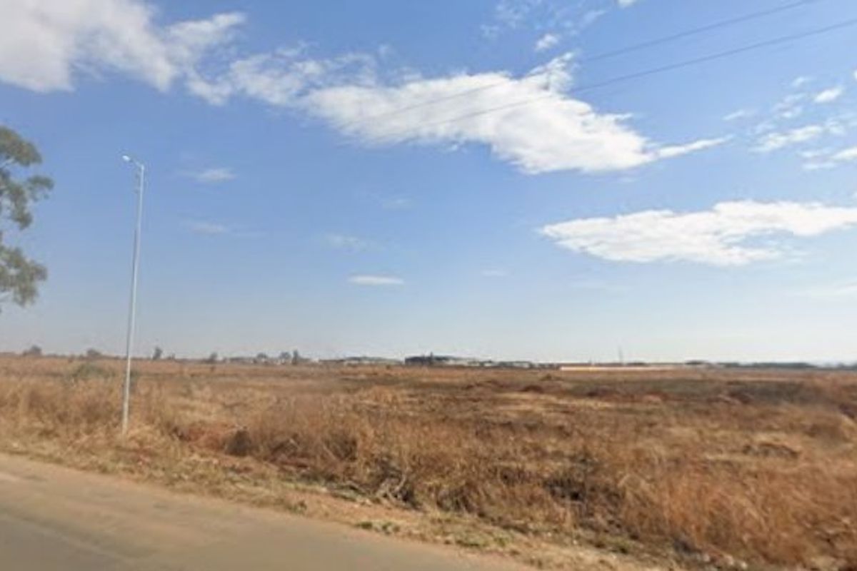 A street-level view of a large, undeveloped residential plot of land adjacent to a paved road with existing street lighting.