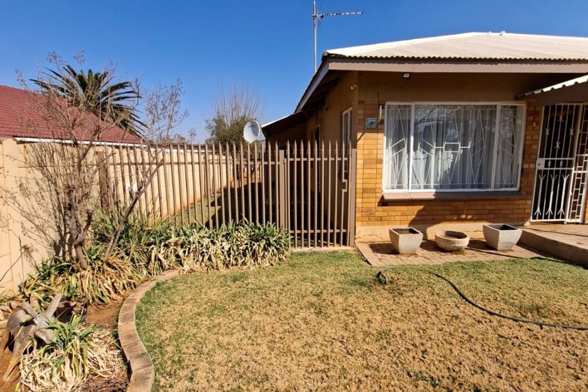 Rear exterior view of a brick house featuring a lawn, garden beds with brick edging, and secure metal fencing.