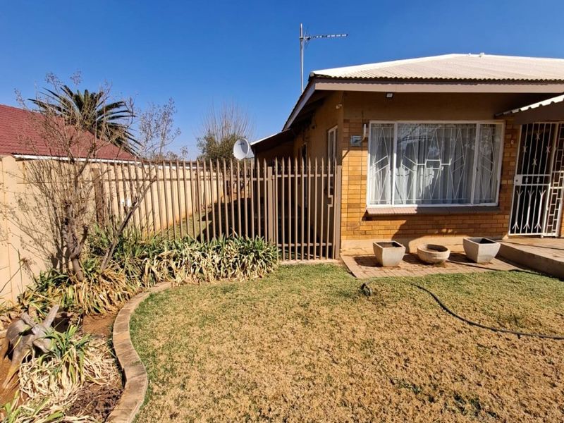 Rear exterior view of a brick house featuring a lawn, garden beds with brick edging, and secure metal fencing.