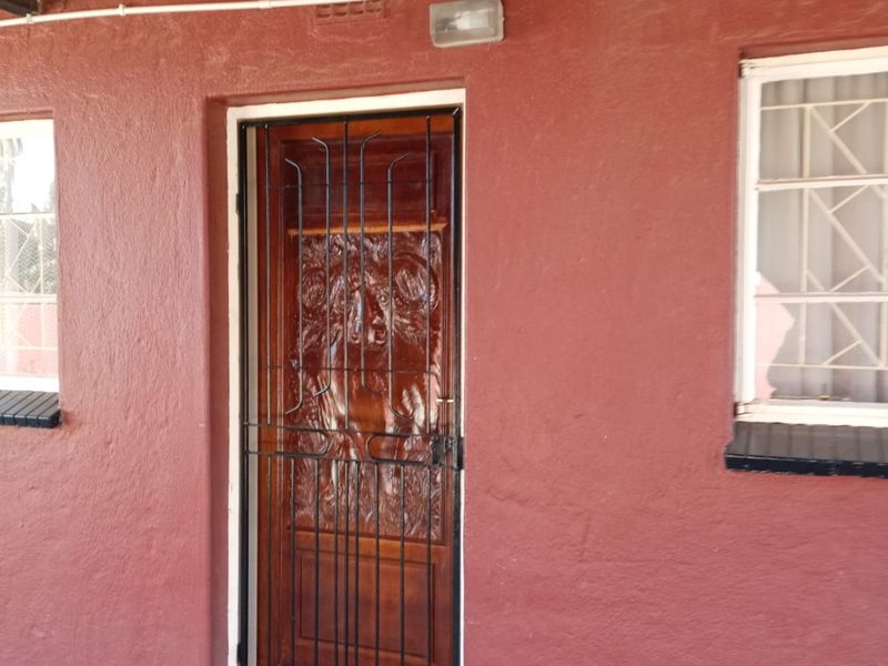 Covered patio area featuring a decorative carved wooden door with security gate, corrugated metal roofing, and windows with burglar bars.