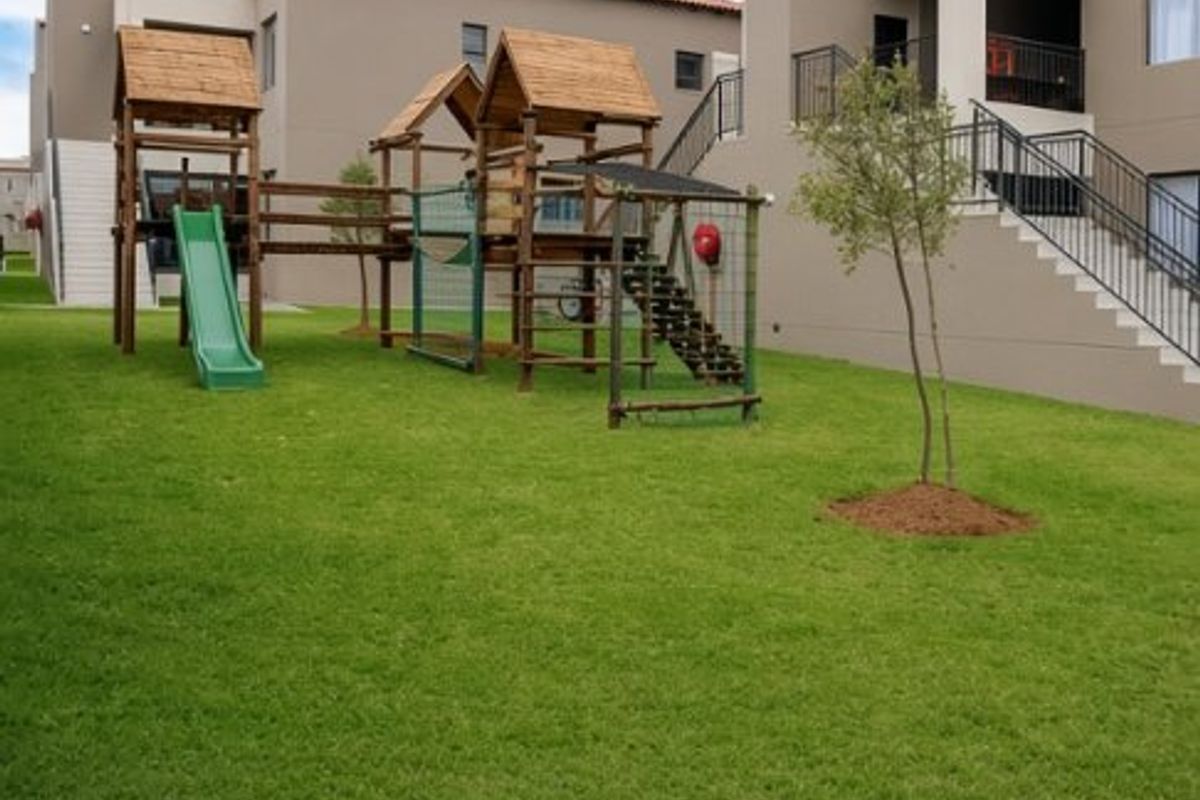 A well-maintained communal garden area featuring a children's play structure, manicured lawn, and access to apartment building balconies.