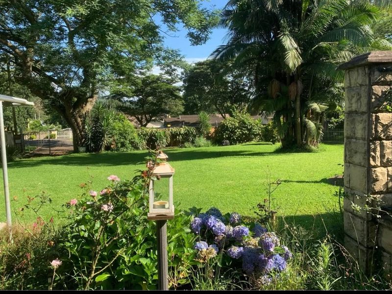 A well-maintained, lush green garden featuring mature trees, established landscaping, and a stone pillar boundary element.