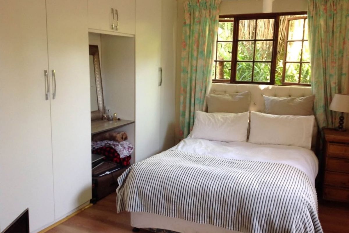 A cozy bedroom featuring built-in cupboards, laminate flooring, and timber-framed windows overlooking a garden area.