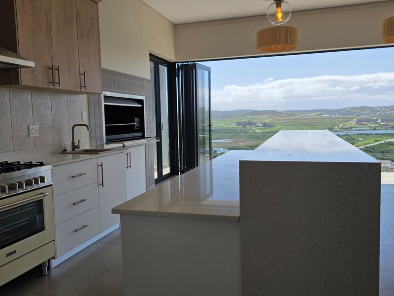 Modern open-plan kitchen featuring stone countertops, a large island, built-in braai, and folding doors opening to expansive landscape views.