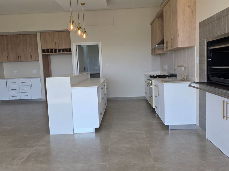 Modern open-plan kitchen featuring sleek white cabinetry, a central island, built-in appliances, and a dedicated indoor braai area with tiled flooring throughout.