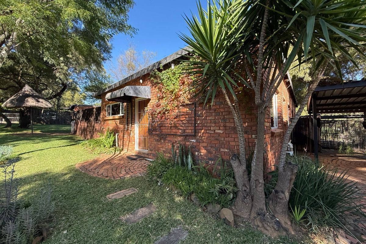 Rear view of the face brick house featuring a paved patio area, security entrance, and adjacent carport set within a landscaped garden.